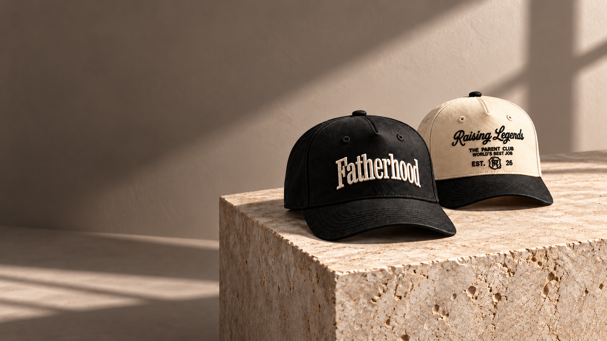 A black Fatherhood cap and cream Raising Legends cap with a black brim resting on sunlit limestone